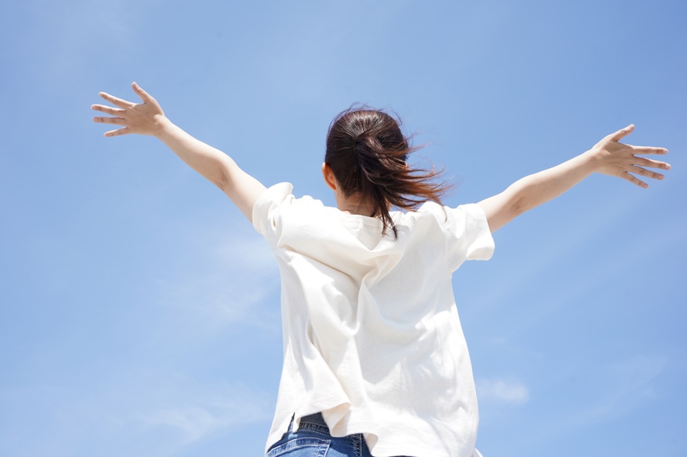 青空に向かって両手を広げて深呼吸する女性の後ろ姿